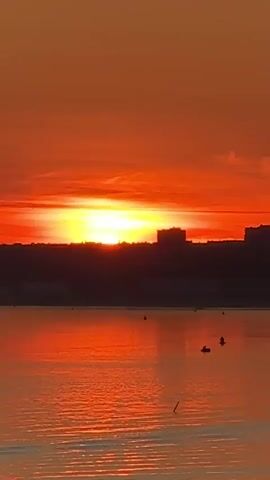 The video shows a sunset over a body of water, likely a lake or river. The sky is a vibrant orange and yellow, with the sun prominently visible just above the horizon, casting a warm glow across the scene. Silhouetted against the bright sky are buildings and trees forming a distant shoreline. Several small objects, possibly boats or buoys, are scattered on the water. The reflection of the sunset shimmers on the water's surface, creating streaks of red and orange. The overall atmosphere is serene and peaceful, with the setting sun dominating the visual frame.