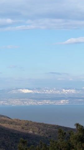 The video shows a distant view of a city across a body of water, with snow-capped mountains in the background. The sky is blue with scattered clouds. The foreground includes trees and vegetation on a hillside. A single ship is visible on the water.