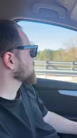 The video shows a man with a beard driving a car on a highway on a sunny day. The camera is inside the car, filming from the passenger seat. The man is wearing sunglasses and a black shirt. Outside the car window, there is a landscape of mountains and trees. The mountains are a light gray color and have jagged peaks. The trees are green and lush. The sky is a clear blue. The car is moving at a moderate speed.