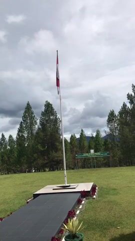 The video shows a national flag of Indonesia flying atop a white pole in a lush outdoor setting. The flag is red and white, and it is positioned centrally in the frame. Below the flagpole is a rectangular platform with black tiles leading up to it, bordered by red flowers and a potted plant. The background is filled with green trees, and a sign is visible in the distance. The sky is overcast with gray clouds. The overall impression is of a solemn or commemorative site in a natural environment.