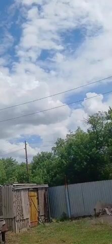 A vertical video opens with a view of a partly cloudy sky above a line of trees. A wooden building with a white foundation is visible in the foreground. Power lines crisscross the frame. The camera pans upwards, revealing more of the sky with cumulus clouds interspersed with blue sky. The camera continues to pan up and to the right, showing the roof of the wooden building and a window. A utility pole is visible behind the building. The camera continues its upward and rightward pan, showing more of the sky and trees in the distance. The video ends with a view of the sky and trees.