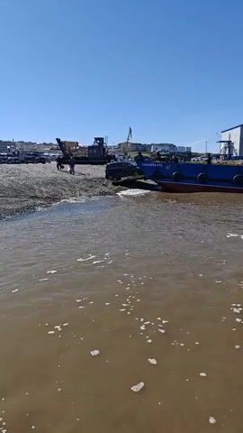 A drone shot moves over murky water towards a blue barge moored near a gravel shoreline. A smaller red barge is partially submerged beneath the blue barge. On the shore, a crane and other industrial equipment are visible, along with a few people. The sky is clear and blue.