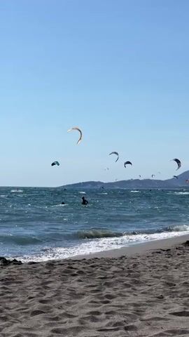 A vertical, eye-level shot captures a beach scene on a clear, sunny day. The foreground is dominated by a sandy beach, with rippled sand textures. The ocean stretches out to the horizon, with choppy, blue waves breaking near the shore, creating white foam. The sky is a solid, bright blue. Several kitesurfers are visible in the water and sky, with their colorful kites dotting the scene. Two people are standing in the shallow water near the shore. In the background, a low, green mountain range is visible against the horizon. The overall atmosphere is one of a breezy, active day at the beach.
