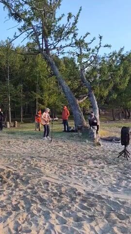 A group of people are gathered on a sandy beach near a forest, with a lone musician playing an acoustic guitar and singing into a microphone. The musician, wearing a plaid shirt and jeans, stands center frame, while several other individuals observe. To the left, two people stand near the edge of the forest. To the right, two people are seen near a large tree, one appearing to be leaning against it. A speaker sits on the ground to the far right. The background consists of a dense forest under a clear sky. The overall scene suggests a casual outdoor gathering or performance.