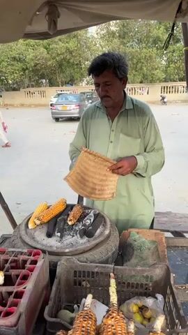 The media file shows a man in Saddar Town, Pakistan, preparing corn on a charcoal grill. He is wearing a light green kurta and has a mustache. The man is using a woven fan to stoke the embers and a metal skewer to turn the corn cobs, which are arranged on the grill.

The scene is an outdoor street food stall, likely operating during the day given the bright, natural lighting. The weather appears to be clear, with no visible clouds. Behind the stall, several cars are parked on a paved surface, and a low wall with vegetation beyond it suggests an urban environment.

In the foreground of the stall, there are crates containing what appear to be drink bottles and more corn on the cob, some of which are already roasted and ready for sale. There are also some fruits, possibly bananas, in a plastic bag. The overall impression is of a typical street-side vendor in a busy area, offering a popular local snack. The man appears focused on his task, with a neutral expression. The image captures a moment of daily commerce and food preparation in a Pakistani setting. There is no visible text in the media file.