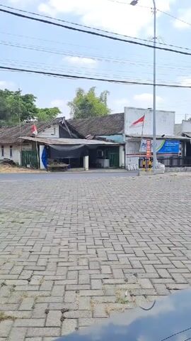 A truck is seen driving past a row of shops. The shops are built in a simple, traditional style with tiled roofs. One of the shops has a red and white flag displayed outside. The street is paved with cobblestones. Overhead, power lines crisscross the sky, and a streetlamp stands on the right side of the frame. The sky is partly cloudy. The video appears to be shot from a slightly low angle, possibly from inside a vehicle.
