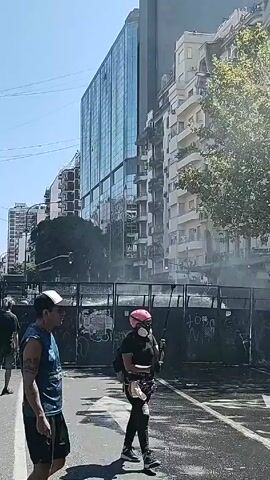 The video shows a street scene with people walking near a barrier. There are several individuals visible: one wearing a pink respirator, another with a white hardhat and plaid shirt, and others wearing casual clothing. The street and barrier appear wet, suggesting recent water application. The barrier itself is adorned with graffiti. In the background, there are multiple buildings, including a modern glass-fronted high-rise and more traditional apartment buildings. The weather seems sunny.