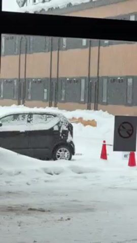 The video shows a snowy scene viewed through a window. A black car is parked in the snow, with a layer of snow covering its roof and parts of its body. Behind the car, there's a building with multiple windows. Orange traffic cones and a sign with a crossed-out cigarette are visible next to the car. Snow is falling.
