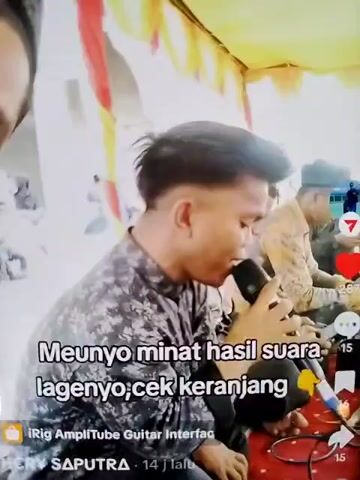A man is singing into a microphone under a yellow and red tent. He is wearing a patterned shirt and has a modern haircut. Another man sits to his right in a tan jacket and hat. The text on the screen reads “Meunyo minat hasil suara lagenyo, cek keranjang” followed by “iRig AmpliTube Guitar Interface”. The video has social media icons for likes, comments, and shares. The environment seems to be an outdoor event.