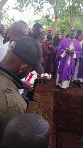 The video shows a burial ceremony. A crowd of people stands around a freshly dug grave. A coffin rests at the bottom of the grave. A priest in a purple robe holds a book and stands near the edge of the grave. Several people hold flowers. The earth around the grave is red. The sky is overcast, and some trees are visible in the background. People appear solemn.