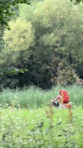A person with a large red backpack is seen cycling through a field of tall grass and wildflowers. The person is wearing a red backpack and light-colored shorts. They are moving from left to right across the frame. The background consists of dense green trees. The video is shot from a distance and is slightly blurry.