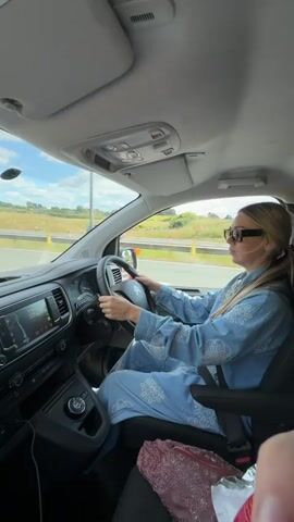 A woman is driving a white van. She is wearing a light blue patterned outfit and sunglasses. She is looking forward and holding the steering wheel. The van is moving on a highway. The passenger side of the van is visible. There is a pink bag on the passenger seat. The sky is cloudy.