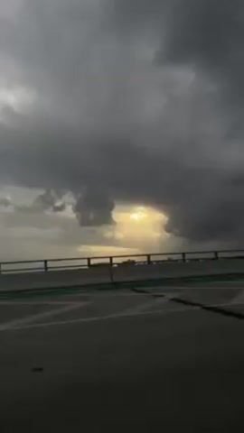 The video shows a dark blue car driving on an elevated road or bridge on a cloudy day. The sun is trying to break through the heavy clouds. The road has white markings and a green railing. A street lamp is visible in the background.