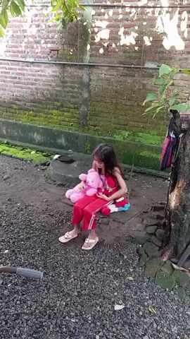 In a medium shot taken from eye level, a young girl sits on a swing outdoors with two stuffed animals. The girl has long dark hair and is wearing a pink tank top and red pants with a white stripe down the side. She is wearing tan-colored sandals. She is holding a pink stuffed teddy bear in her left arm and a small stuffed doll in her right arm.

The swing is made of wood and hangs from two metal chains, suspended from a tree branch. The ground beneath the swing is covered in small dark gray gravel. A section of a bicycle handlebar is visible in the lower left corner of the frame.

Behind the swing is a brick wall covered with moss and some green plants. A gray concrete base runs along the bottom of the wall. To the right of the swing, the trunk of a tree is visible. A pink and black bag hangs from the tree.

The lighting is bright and sunny, casting shadows from the tree branches and leaves above. Some patches of grass are seen on the ground near the wall and around the base of the tree. The background suggests a backyard or garden setting.