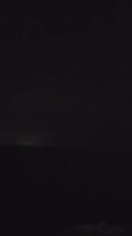 The video captures a nighttime scene over a body of water, likely the ocean, under a dark and cloudy sky. There are intermittent flashes of lightning illuminating the clouds from within, suggesting a thunderstorm in progress. The water's surface is dark and somewhat rippled. No people or man-made objects are clearly visible, though a faint light, possibly a distant buoy or boat light, can be seen on the horizon in the second half of the clip. The overall atmosphere is dramatic and moody due to the storm and the darkness.