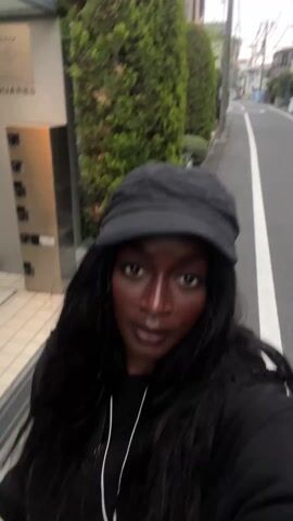 A person with long dark hair and dark skin wearing a black hoodie with white drawstrings and a black cap walks along the sidewalk next to a narrow street. The person holds their phone in front of them and looks at the camera.  Green hedges and a building are visible along the side of the road.