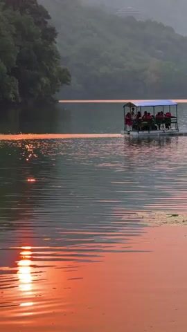 A serene wide shot captures a boat gently gliding across a calm lake during sunset or sunrise. The water reflects vibrant orange and red hues from the sky, creating a striking contrast with the cooler tones of the distant mountains. A small, flat-bottomed boat with a blue canopy shelters several people, who appear to be enjoying a peaceful ride. The boat is moving from left to right, leaving a subtle wake. Lush green mountains, partially shrouded in a soft haze, form the backdrop, with a few buildings visible on one of the distant slopes. The overall scene conveys a sense of tranquility and natural beauty, characteristic of a mountainous lake region like Pokhara, Nepal. There is no visible text.