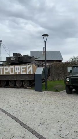 The video shows military vehicles, likely on display. The video starts with an angled shot of an olive green military vehicle covered with netting and parked on a paved surface. To the left are tanks and a black sign. Behind the vehicle is grass, a fence, and a building with a gray, slanted roof. The camera pans to the right to reveal a small evergreen tree and then a large, green helicopter. A man wearing a hat, a gray-and-white jacket, and dark pants walks by the helicopter as it is filmed. The sky is cloudy.