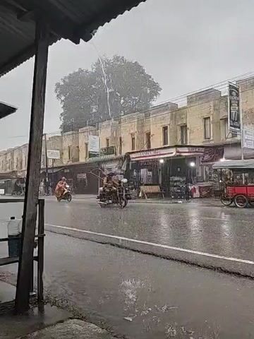 The video shows a street scene during heavy rain. The view is from under a shelter, likely a storefront awning. Across the street are several buildings with awnings over storefronts. A large tree is visible further down the street. Rain is falling heavily, and the road is wet with visible puddles. Several motorbikes are seen driving on the road. One red cart is parked near the shops. The sky is overcast and gray, contributing to the dreary atmosphere.