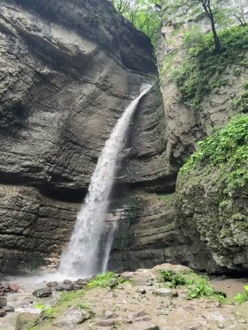 This is a video of a waterfall cascading down a rocky cliff face. The water is a light white, creating a misty spray as it falls. The cliff is made of layered rock, with shades of grey and brown. Green foliage grows on the top and sides of the cliff, and some green plants are visible at the base of the waterfall. The ground in front of the waterfall is rocky and appears to be wet, with some small pools of water. The overall environment suggests a natural, lush setting. There are no people or animals visible in the video. The footage appears to be shot from a slightly elevated position looking up at the waterfall.
