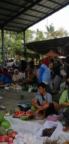The video shows a busy outdoor market scene, captured with a vertical orientation. The market appears to be set up under a covered structure with a corrugated metal roof, and the background shows lush green trees and vegetation, suggesting an outdoor, possibly tropical, setting. The ground is a concrete or dirt surface.

In the foreground, on the right side, there are several stalls displaying fresh produce. Prominently featured are a large pile of watermelons, some cut in half to reveal the red flesh, and a bunch of red grapes. There are also other fruits and vegetables, including what appear to be dragon fruits and smaller green fruits that might be limes or small melons. A young man wearing a gray t-shirt is crouched on the ground, facing towards the right, seemingly attending to his stall.

The market is filled with many people, predominantly women, dressed in various colorful clothing and headscarves. They are gathered around different stalls, interacting with vendors, examining produce, and making purchases. The overall impression is one of a lively and active marketplace.

The perspective of the camera is at eye level with the people in the market. The lighting suggests it is daytime, possibly with overcast conditions given the diffused light. The video is relatively still, with only slight camera movements, allowing the viewer to take in the details of the market. The arrangement of the stalls and the presence of customers suggest a typical local market setting.