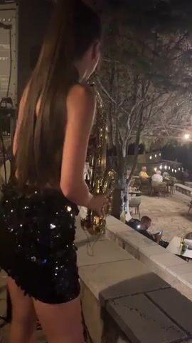 In this media file, a young woman with a high ponytail, wearing a sparkly black mini-dress, plays a tenor saxophone. She is positioned outdoors on a raised stone terrace, which appears to be part of a restaurant or outdoor venue. In the background, several tables with diners are visible, suggesting a social gathering or event. Trees with lush foliage are behind the woman, and strings of lights are strung across the scene, indicating it is evening. The lighting suggests it is dusk or nighttime, and the atmosphere seems relaxed. A man is seated at a table in the middle ground, looking at his phone. The woman's body language indicates she is focused on her performance. There is some visible text on a piece of equipment to the left, appearing to be a speaker or sound system, with the brand name "FBT" faintly discernible. The overall impression is of a live musical performance taking place at an outdoor dining establishment.