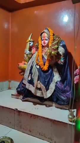 A statue of a goddess, adorned with orange flower garlands and rich dark blue and gold robes, sits on a raised platform within a dimly lit chamber. The statue has a serene expression, with red markings on its forehead and cheeks. To the left of the goddess, a tiger statue is partially visible, with the goddess holding a golden weapon in its right hand. The walls of the chamber are painted a warm, orange hue, and the floor appears to be tiled. There is also a glimpse of pink flowers and a small brass vessel in the foreground. The lighting suggests an interior setting, possibly a temple or shrine, with light emanating from above.