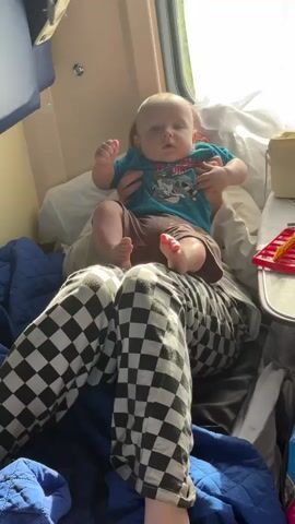 A baby sits on someone's lap, facing forward, with their legs spread apart. The baby is wearing a blue t-shirt with a graphic design and brown shorts. The person holding the baby is wearing black and white checkered pants. The scene appears to be inside a train compartment, with a window on the right side and bedding visible. The lighting suggests it is daytime.