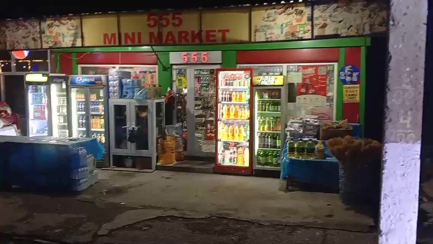 The video shows the exterior of a mini market at night. The mini market has a sign that says "555 MINI MARKET." There are several refrigerators outside, filled with drinks. There are also displays of water bottles, cooking oil, and snacks. The ground in front of the store is wet. The video is taken from a low angle and pans slightly to the right. The lighting is dim, with the refrigerators and signs providing the main sources of illumination.