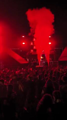 The video shows a concert at night. A large crowd of people are gathered in front of a stage. Red lights and smoke effects illuminate the stage and the sky. The stage features multiple light panels and some performers. The overall atmosphere appears to be energetic and exciting.