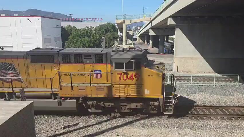 The video shows a train comprised of at least three locomotives moving on a track under an overpass. The first locomotive is yellow with an American flag painted on its side and the number 7049. The second locomotive is also yellow and displays the number 4830. The third locomotive, yellow with the number 5563, is visible at the end of the sequence. The train has graffiti on at least two of the locomotives, including the word "ENEMY" and "REBEL". The environment appears to be an industrial or urban setting with a building and trees visible in the background, along with a concrete overpass. The lighting suggests it is daytime with a clear sky.