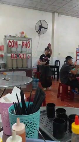 The video captures the interior of a modest Vietnamese restaurant. A young woman in a black t-shirt and patterned pants walks towards the back of the room, carrying a small bowl and a tray. Another man is seated on a red stool, eating. The restaurant has tiled floors and white walls with some minor stains.  A black fan is mounted high on the wall.  A glass display case labeled "COM - PHO HA NOI" showcases stacks of bowls.  In the foreground, a table displays containers filled with chopsticks, napkins, and condiments.  Several black cups are arranged on a tray.