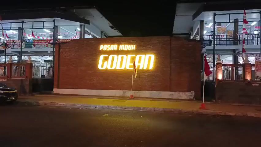 The video shows a brick wall with illuminated text that reads "PASAR INDUK GODEAN". The wall is lit by yellow lights. There are buildings with glass windows on either side of the wall, which appear to be market stalls or shops. Indonesian flags are visible on poles in front of the buildings. A street with asphalt pavement is in the foreground, and a dark-colored car is parked on the left side of the street. The scene is illuminated by artificial lights, indicating it is nighttime.