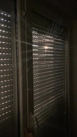 The video shows a close-up view of a window with closed horizontal blinds, seen from indoors. Light from outside shines through the gaps in the blinds, creating bright lines on the surface. The room is otherwise dark. The window has a white frame and a handle on the right side. The camera is static throughout the video.