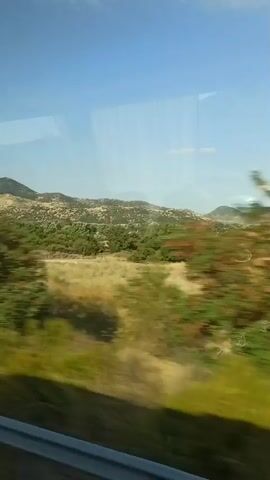 The video captures a view from a moving vehicle, likely a train, looking out a window. The scene outside is a landscape with hills in the distance covered in sparse vegetation and trees. In the middle ground, there are more dense trees and fields with dry grass or bushes. A dirt road is visible with what appears to be a green tractor and a dark vehicle on it. The sky is clear and blue. The motion blur indicates the rapid movement of the vehicle.