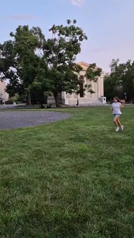 A young girl with long brown hair runs into frame from the right and stops at an Adirondack chair in a grassy area. She turns and sits down in the chair. The background consists of trees and a large building.