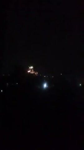 The video shows a dark night scene. In the distance, there are clusters of yellow and white lights that intermittently appear to be fireworks. The foreground is mostly black, with a few blurry dark shapes, possibly trees or buildings. A bright white light appears stationary on the ground in the middle distance. There are other, more faint, white lights also visible in the distance, presumably from buildings or streetlights. The sounds of fireworks are audible throughout.