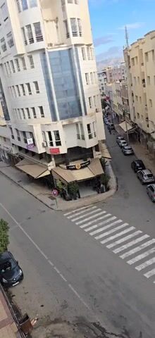 The video shows an aerial view of a street intersection in a city.  Several buildings surround the intersection. There are parked cars along the sides of the streets. A pedestrian crosswalk spans one of the streets.  People are walking across the street and riding bikes. A cafe called "Cafe Malaki" is located on a corner. There is a sign for a photo studio, "Labo Photo Colisee."  The scene appears to be taken during daylight hours.