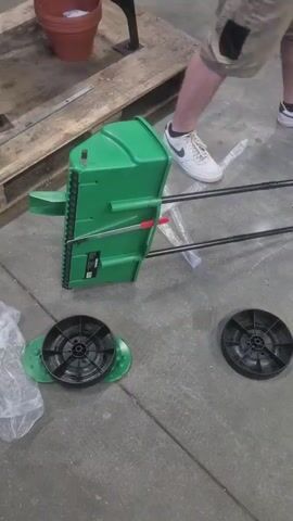 In an indoor setting resembling a warehouse or workshop, a person wearing khaki shorts, white athletic shoes, and a white t-shirt is standing on a concrete floor. To their left, a green piece of equipment, possibly a lawn spreader or sweeper, is partially visible, lying on its side. Attached to it are two black metal rods, likely handles. In front of the green equipment, two black circular wheels with intricate spokes are laid out on the floor. One wheel is positioned on a green base plate, while the other sits independently. A clear plastic bag, crumpled and partially empty, lies to the left of the wheels. Further in the background, a wooden pallet supports a terracotta pot. A dark metal object, possibly part of a table or stand, is also visible. The lighting suggests it is daytime, but there are no external weather cues. The overall scene appears to be a workspace where an item is being assembled or prepared for use.