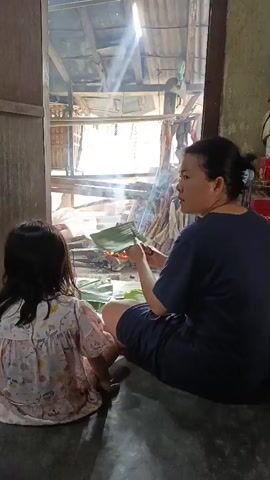 A woman and a young child are sitting on the floor in front of an open doorway. Sunlight streams through the doorway, illuminating smoke rising from a small fire pit. The woman, facing away from the camera, is holding a green leaf in her right hand and appears to be speaking to the child. The child, also facing away from the camera, is wearing a patterned dress and is looking towards the fire. The environment appears to be rustic, with visible wooden structures and hanging fabric in the background.