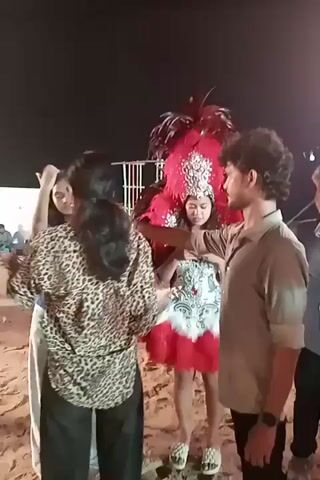 A young woman in a carnival costume is holding a phone and looking at it while surrounded by several other people in what appears to be an outdoor setting at night. The woman is wearing a red and white costume with large red feathers on her back and a feathered headpiece. The ground is sandy. A man with curly hair and a light-colored shirt stands to her right, looking at her. Another woman in a leopard-print top is visible to her left, walking away. In the background, there are some structures made of bamboo poles and string lights, suggesting a festive or event atmosphere.