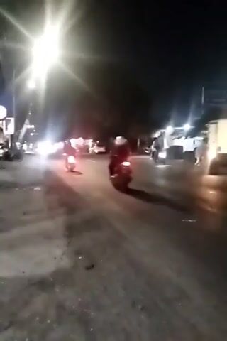 A night-time video captures a street scene with several motorcycles and a truck. Bright streetlights illuminate the road, creating lens flares and hazy effects. On the left side of the road, a building with red and white banners is visible. Several motorcycles are seen moving in both directions, their headlights shining brightly. A white truck is parked on the right side of the road. The road surface appears worn and somewhat rough.