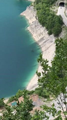 A high-angle shot shows a winding road alongside a body of water. The road has a tunnel at one end, and a motorcyclist is riding towards it. The water is a deep blue-green. The shoreline is rocky with a small sandy beach. There are trees and vegetation along the side of the road and on the hillside.