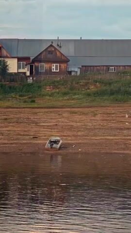 The video captures a serene riverside scene in Vorogovo, Russia, likely during late afternoon or early evening given the soft, warm light and gentle reflections on the water. The foreground is dominated by the rippling surface of a body of water, its surface catching the ambient light and creating a shimmering effect.

Beyond the water, a dry, sandy riverbed stretches out, dotted with a small, weathered boat resting on the exposed ground. A lone figure, likely a person, is seated in the boat, creating a sense of quiet contemplation or perhaps a moment of rest.

The middle ground features a low-lying bank covered in dry grass and dirt, indicative of a low water level. This dry area contrasts with the lush green vegetation that grows further up the bank, forming a vibrant border.

In the background, a collection of rural houses are visible, nestled against a backdrop of trees. The architecture suggests a traditional Russian village setting, with wooden structures and pitched roofs. The houses appear weathered, suggesting age and a lived-in quality. The sky above is a soft blend of blue and hints of pink or orange, reinforcing the impression of a peaceful twilight. The overall mood is tranquil and evokes a sense of rural simplicity and quietude. There is no visible text in the media.