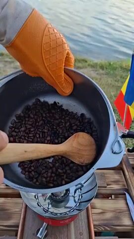 This is a close-up, top-down shot of someone roasting coffee beans. The scene appears to be outdoors, possibly a campsite, with a wooden table in the foreground. A silver camping stove is visible, with a small pot of water or coffee brewing equipment to its right. A person wearing orange oven mitts is holding a metal pot filled with dark brown coffee beans. They are using a wooden spoon to stir the beans, some of which are falling from the spoon back into the pot. A yellow can with a red logo is in the background, as well as a body of water. The lighting suggests it is daytime.