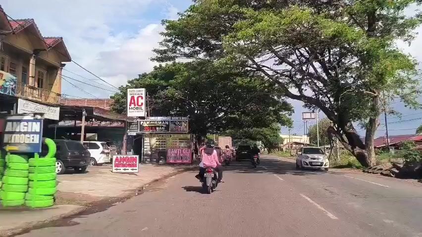 The video shows a view from a car driving down a road on a sunny day. On the left side of the road, there is a small business with a sign that reads "PAK USMAN OLI & BAN". There are several cars parked in front of the business. A person on a scooter is riding down the road in the same direction as the car. There are other cars and scooters on the road as well. The road is surrounded by trees and buildings. Power lines are visible overhead.