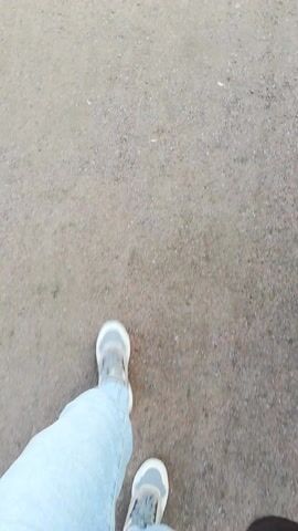This media file captures a first-person perspective of someone walking on a gravel path in Kolpino, Russia. The scene transitions from a close-up of the walker's feet in jeans and sneakers to a wider view of the path as it winds through a park-like setting. Lush green grass borders the path, and mature trees with dense foliage create a shaded canopy in some areas. The sky is visible, showing patches of white clouds against a bright backdrop, suggesting it is daytime, likely late morning or early afternoon, with fair weather.

Further along the path, other individuals are visible, including a person on a bicycle in the distance and what appear to be other park-goers on the grass. The path curves, leading the viewer's eye deeper into the scene. Beyond the park area, glimpses of urban elements emerge. A building with several stories, possibly an apartment complex, is visible on the left side of the frame. A few cars are parked near a fence, and a bus can be seen in the background, indicating the proximity of residential areas and public transportation. The overall atmosphere appears peaceful and casual, typical of a leisurely walk in a park on a pleasant day. No visible text is present in the media file.