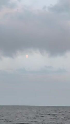 The video shows a sky with clouds over water. The moon is visible through the clouds. The colors of the sky are muted, with shades of gray, white, and hints of pink. The water appears dark and slightly choppy. The scene suggests an outdoor environment, possibly at dusk or dawn.