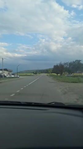 This is a first-person perspective video taken from inside a car driving along a road. The scene is rural, with a paved road stretching ahead, bordered by grassy fields and trees. In the distance, rolling hills can be seen under a partly cloudy sky. The sky is a mix of blue and gray clouds, suggesting it might be late afternoon or early evening, possibly with a storm approaching in the distance. The road has a solid white line marking the center and dashed white lines on the right side. On the left side of the road, a building resembling a small restaurant or shop is visible, with a few parked cars. The overall mood is calm and transitional, as if traveling from one place to another. There are no visible people or spoken words, focusing solely on the journey and the surrounding landscape. The location is identified as Alkhasty, Russia.