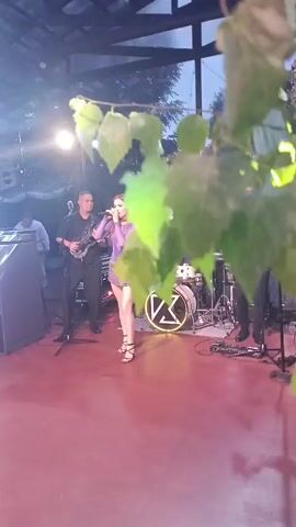 A band performs on an outdoor stage at dusk. A female singer in a purple sequined dress stands center stage, singing into a microphone and gesturing with her arms. To her left, a male guitarist in a black suit plays an electric guitar. To her right, another male musician plays what appears to be a keyboard or synthesizer. In the background, a drummer is seated behind a drum kit with a circular logo featuring the letter "K" in neon light. The stage is covered by a wooden canopy with visible rafters. Several trees and foliage are visible in the background, suggesting an outdoor venue. Lighting on stage includes red spotlights and colorful moving lights that project patterns onto the floor.